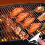 Tenedor BBQ inoxidable asando alitas y carne en parrilla con llamas.