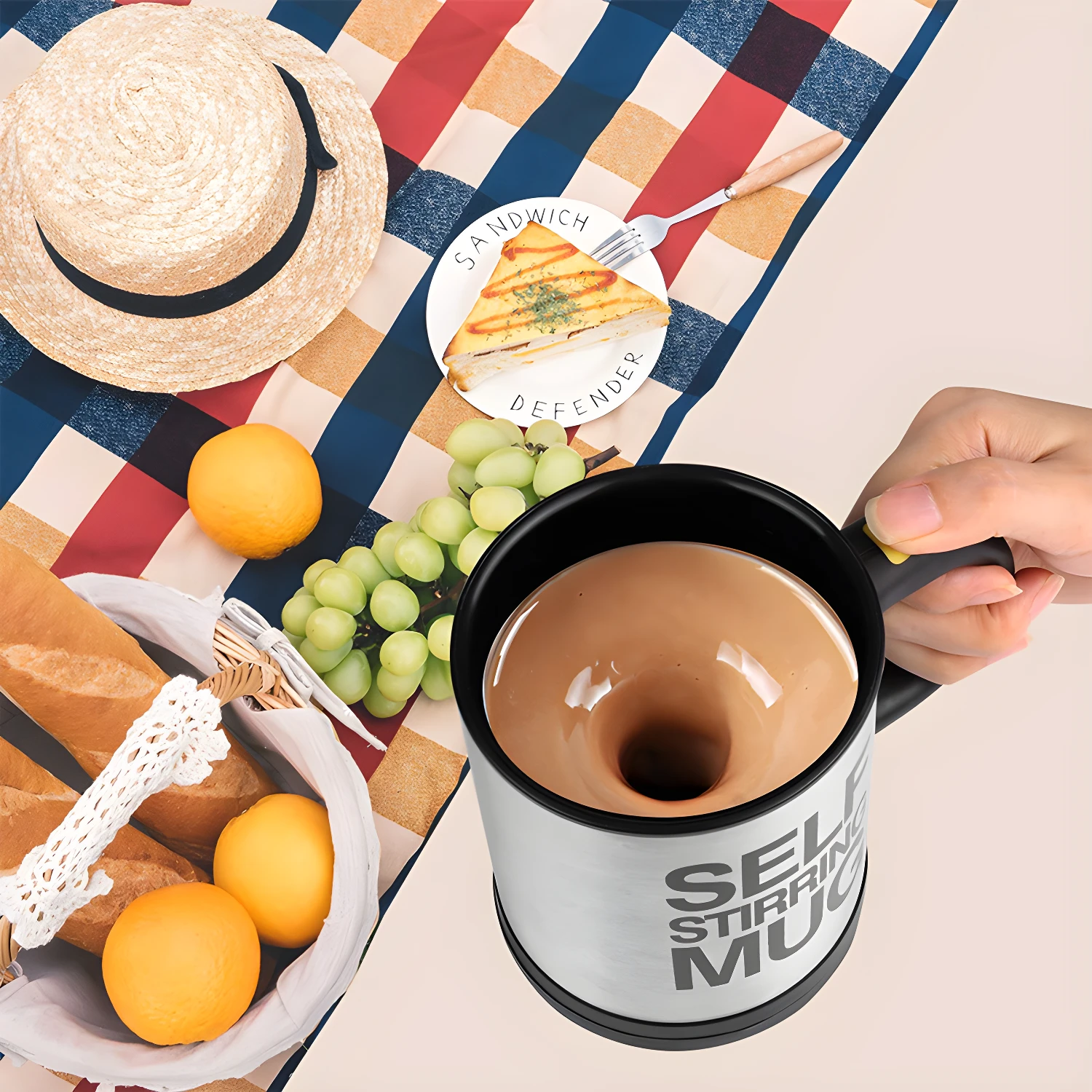 Pocillo mezclador auto-agitador con café, listo para picnic con frutas, sándwich y sombrero.