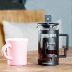Combo Molino Café Y Prensa Francesa: taza rosa y cafetera francesa negra con posos de café.