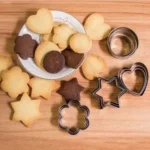 Cortadores de galleta en forma de estrella, corazón y flor, rodeados de galletas horneadas.
