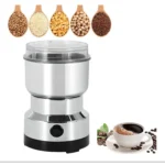 Molino de café eléctrico plateado junto a taza de café, granos y cucharas con ingredientes.