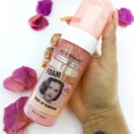 Bote rosado de removedor de maquillaje Kiss Beauty Real Fresh Foam, sostenido en mano, rodeado de pétalos.