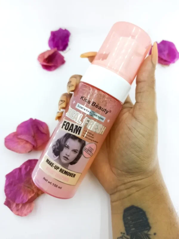 Bote rosado de removedor de maquillaje Kiss Beauty Real Fresh Foam, sostenido en mano, rodeado de pétalos.