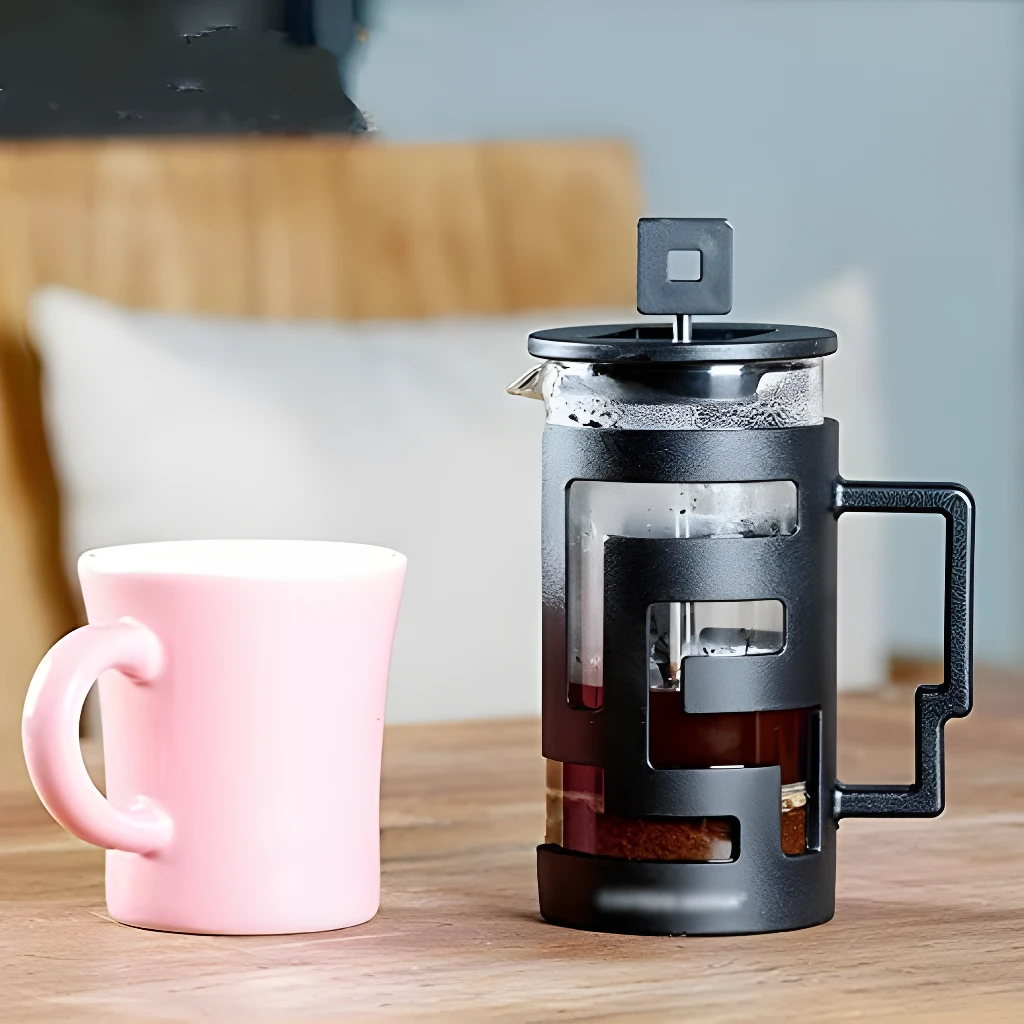 Prensa francesa negra y taza rosa sobre mesa de madera, ideal para café.