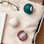 Esferas atrapa-pelos en verde y blanco, con detalles dorados, junto a gafas y libro.