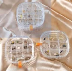 Organizador de joyero transparente con varios compartimentos exhibiendo anillos, aretes y collares dorados.