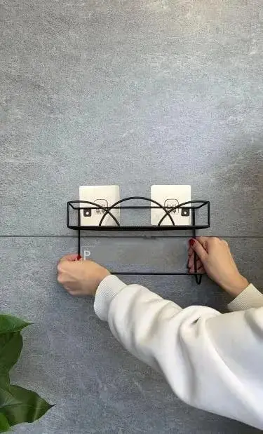 Estante de pared negro con soporte y dos objetos blancos colgando, instalado sobre pared gris.