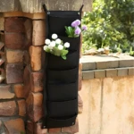 Bolsa para plantar vertical negra con flores blancas y lilas en pared de ladrillo.