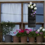 Bolsa para plantar vertical negra con flores blancas y moradas, colgada en ventana con macetas de geranios.