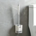 Cepillo de baño blanco y gris para inodoro, montado en pared junto a un inodoro blanco.