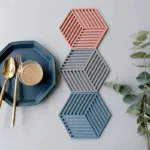 Posavasos de silicona hexagonales apilados en tonos coral, gris y azul, junto a un plato azul y cubiertos dorados.
