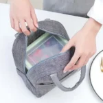 Lonchera térmica gris con contenedor transparente y asas, ideal para llevar alimentos.