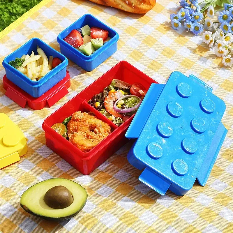 Fiambrera lonchera Lego azul y roja con comida y aguacate sobre mantel a cuadros amarillos.