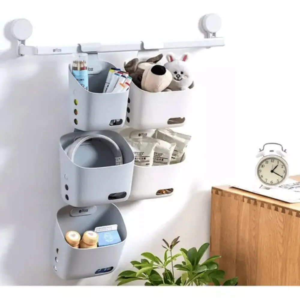 Cesta organizadora colgante gris y blanca con juguetes y artículos diversos, junto a un reloj despertador.