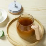 Taza mezcladora eléctrica con asa de madera y bebida marrón sobre bandeja de madera.