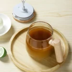 Taza mezcladora eléctrica con bebida marrón y rodaja de limón en bandeja de madera.