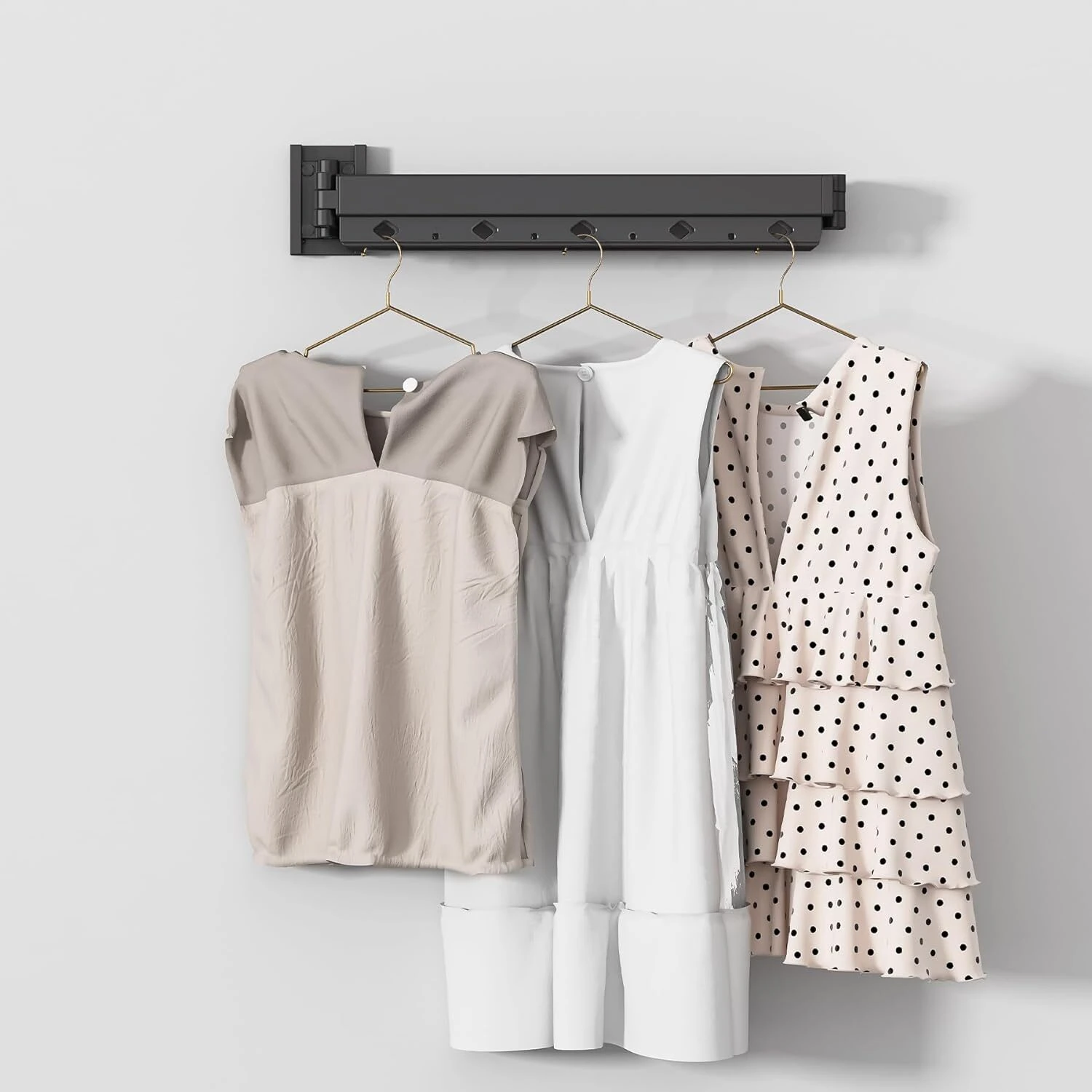 Tendedero plegable de pared con tres vestidos colgados: beige, blanco y estampado de lunares.