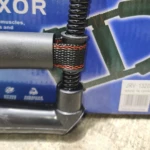 Ejercitador de mano, muñeca y antebrazo con resorte negro y banda de sujeción, sobre caja azul.