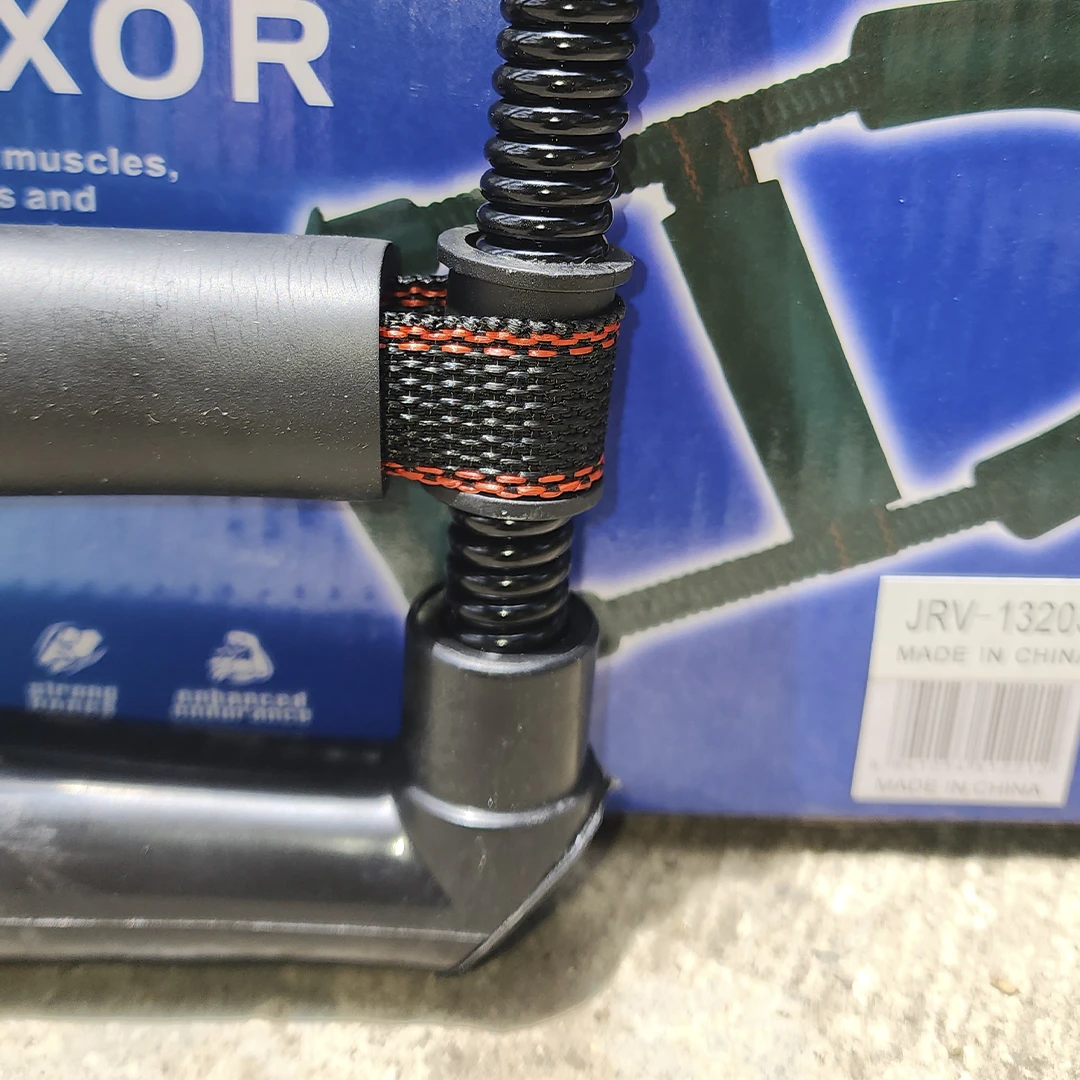 Ejercitador de mano, muñeca y antebrazo con resorte negro y banda de sujeción, sobre caja azul.