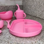 Vajilla de silicona para bebé en color rosa: plato con divisiones, vaso con popote, tazón y cubiertos.