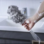 Cepillo corporal de ducha con esponja gris, genera espuma para limpieza profunda.