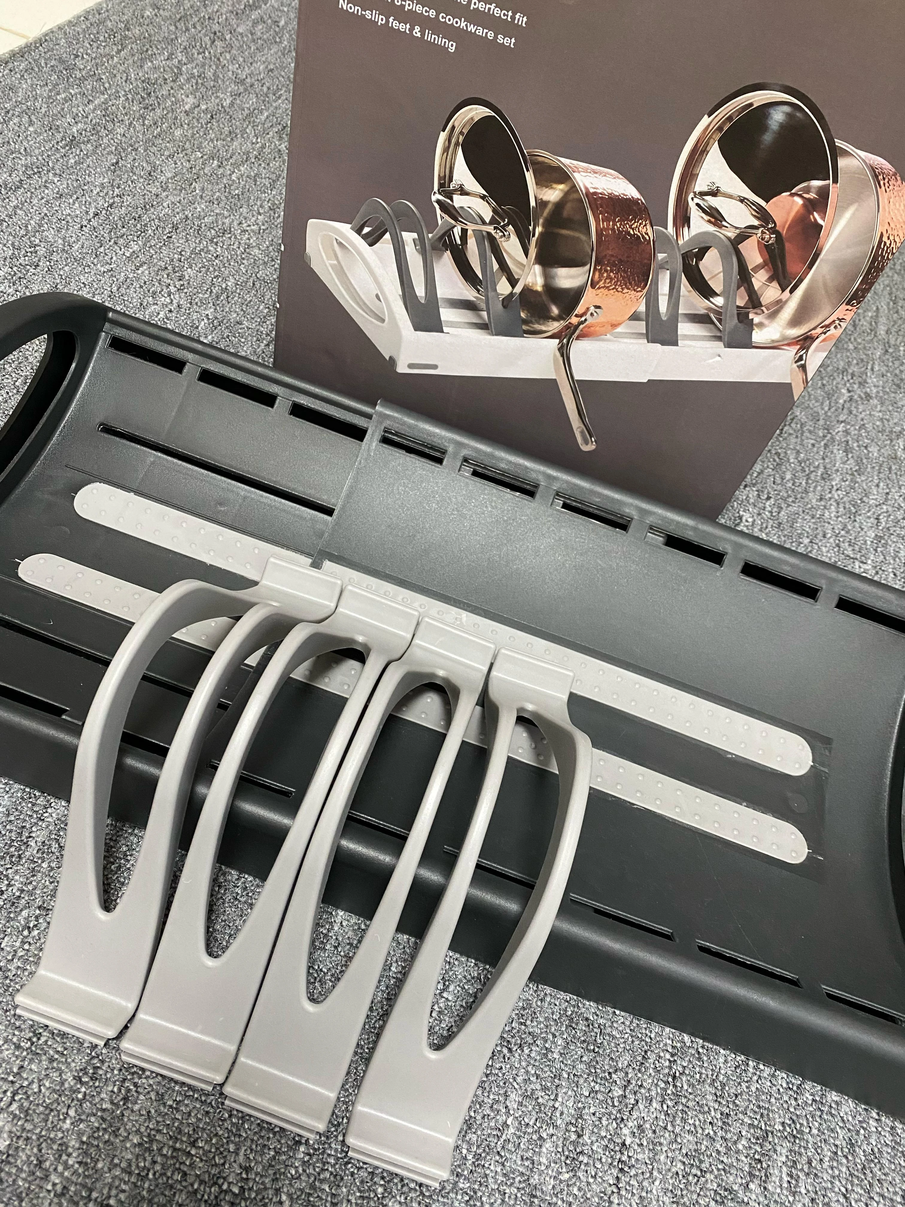 Organizador de ollas ajustable en gris sobre alfombra, con ollas de cobre en la caja.