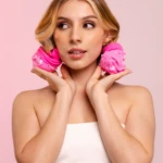 Mujer joven con medias rizadoras rosas en pelo rubio, mirada coqueta sobre fondo rosa.