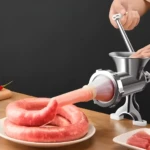 Molino de carne manual procesa carne para salchichas con boquilla y plato blanco.