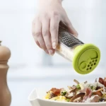 Rallador manual con contenedor esparciendo queso sobre pasta fresca con jamón y champiñones.