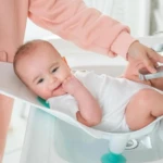 Bebé sonriente en soporte de bañera azul y blanco, con mano ajustando grifo.