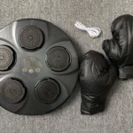 Máquina de boxeo portátil negra con guantes y cable USB sobre fondo de tela gris.