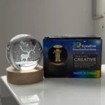 Lámpara esfera de cristal 3D con diseño de ciervo y estrellas, junto a su caja.