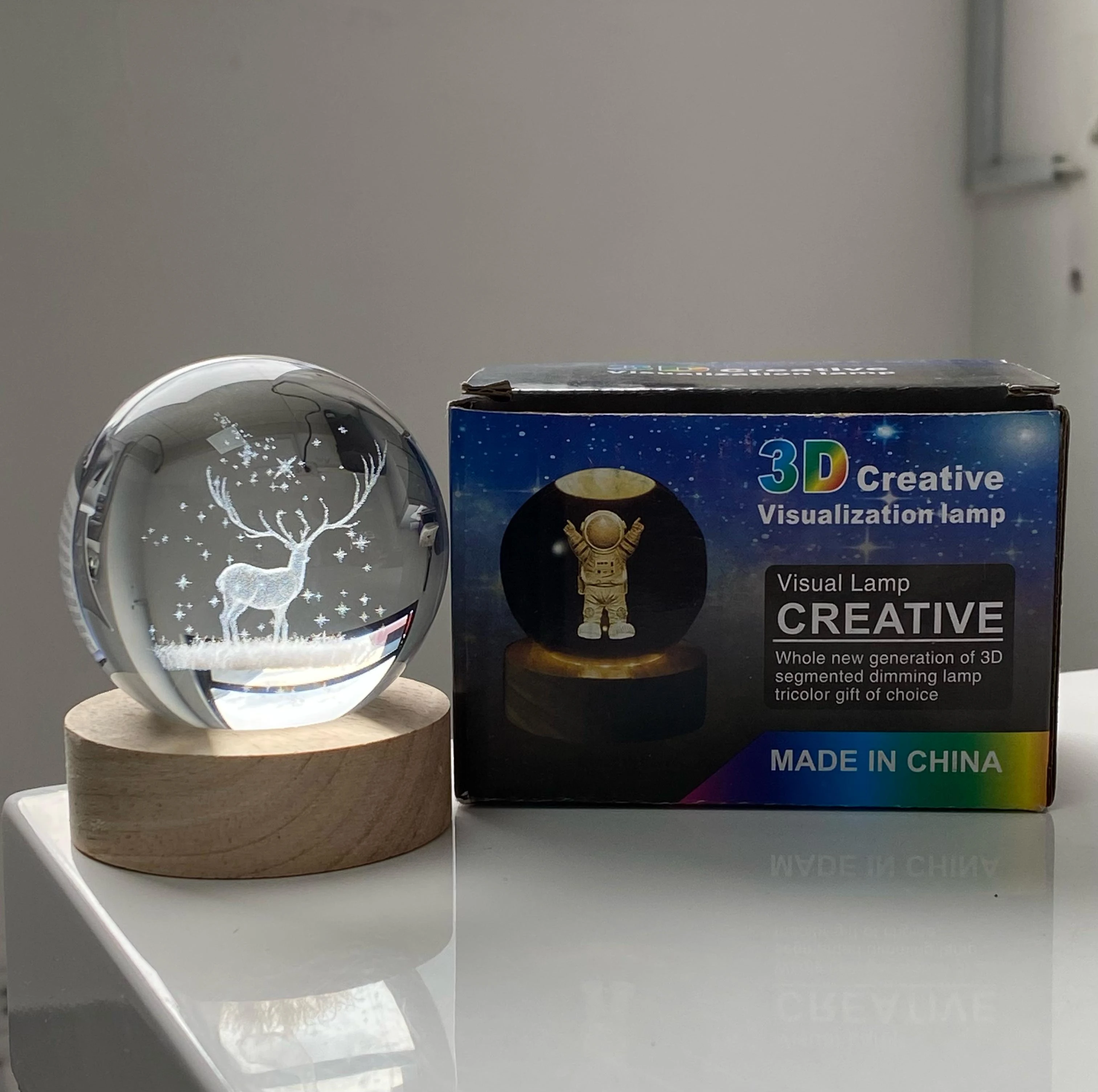 Lámpara esfera de cristal 3D con diseño de ciervo y estrellas, junto a su caja.
