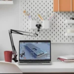 Brazo flexible negro para sujetar celular, tablet o cámara web sobre un escritorio blanco junto a un laptop y una taza roja.