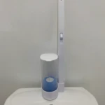 Cepillo limpiador de inodoro desechable blanco, con vela azul en soporte, sobre cisterna de inodoro blanco.