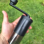 Moledor de café manual negro con recipiente transparente sostenido en mano sobre césped.