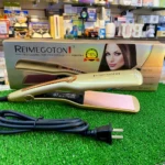 Plancha de cabello Reimegoton dorada con placas de cerámica rosa, sobre su caja original en un mostrador de tienda con césped artificial y cable de red.