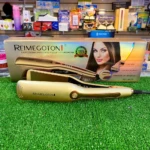 Plancha alisadora Reimegoton dorada modelo RE-9677 frente a su caja, exhibida sobre césped artificial en un establecimiento comercial.