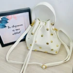 Bolso carriel blanco tipo saco con tachuelas doradas, cordón ajustable, asas y correa larga, junto a cuadro decorativo.