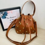 Bolso tipo carriel color café caramelo con taches dorados, estilo saco ajustable, junto a cuadro decorativo.