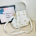 Bolso tipo carriel blanco de cuero sintético con taches dorados, cordón ajustable y correa larga, junto a un cuadro con frase motivacional.