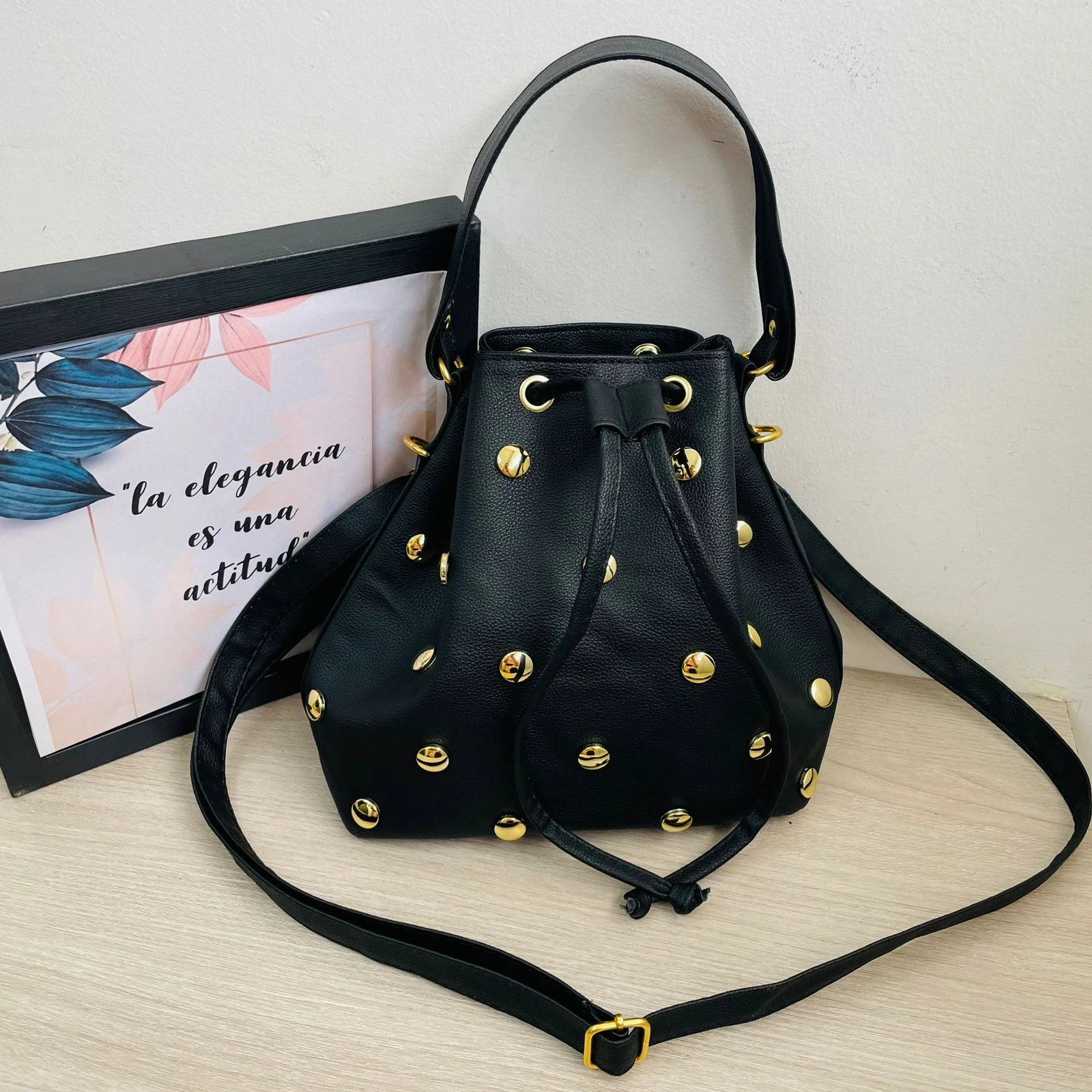 Bolso tipo carriel negro de cuero con taches dorados y cordón ajustable, junto a cuadro decorativo.