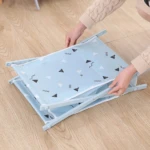 Manos plegando canasta organizadora de tela azul claro con triángulos blancos y negros sobre piso de madera clara.