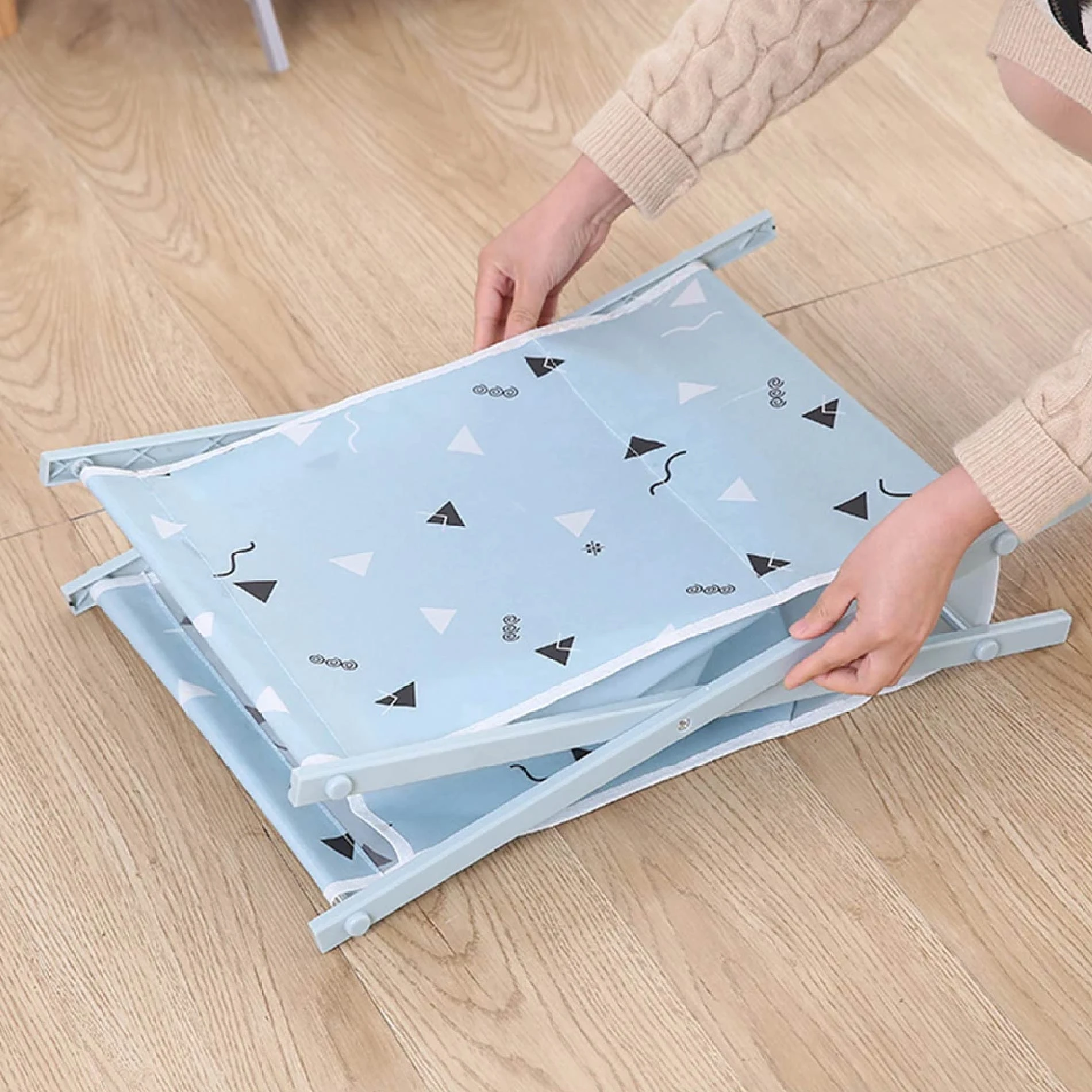 Manos plegando canasta organizadora de tela azul claro con triángulos blancos y negros sobre piso de madera clara.