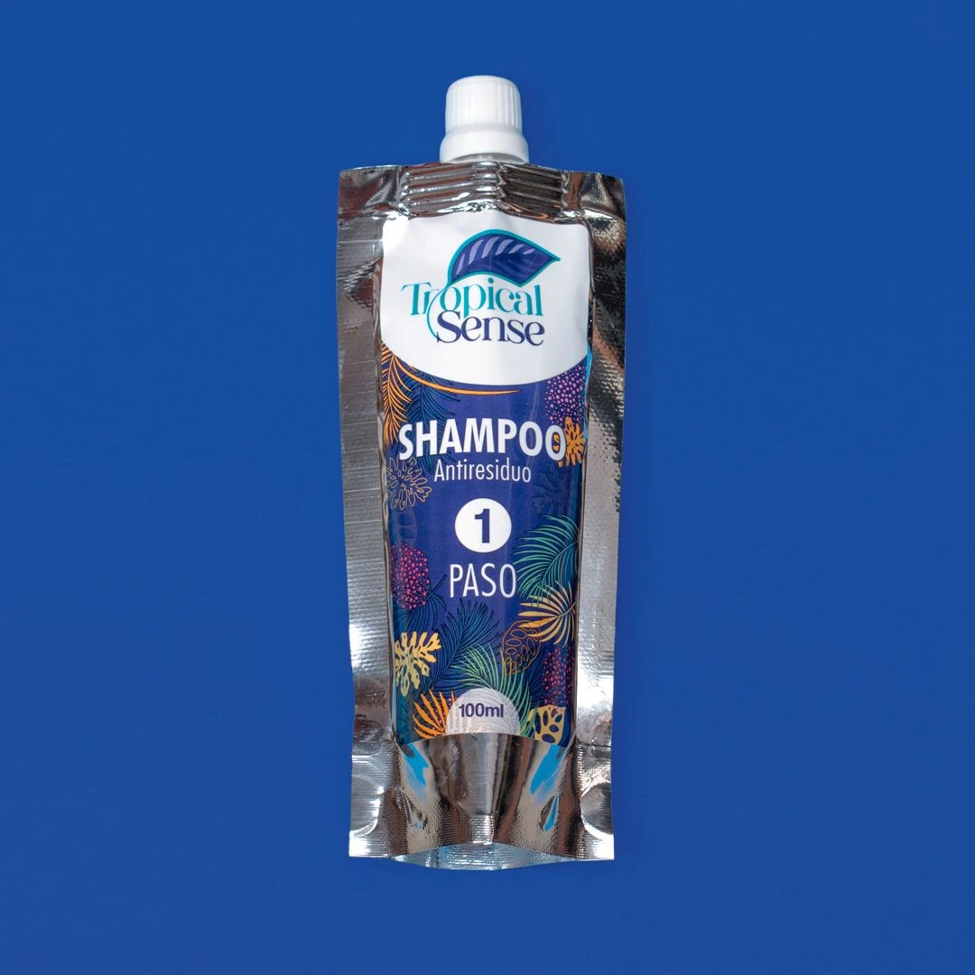Envase flexible plateado de 100 ml con champú antiresiduos Tropical Sense, paso 1 del tratamiento, sobre fondo azul sólido.
