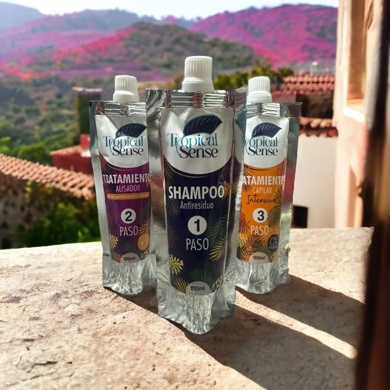 Kit de tres sobres de 100ml para alisado capilar Tropical Sense ordenados por pasos sobre una superficie de piedra con paisaje de montaña de fondo.