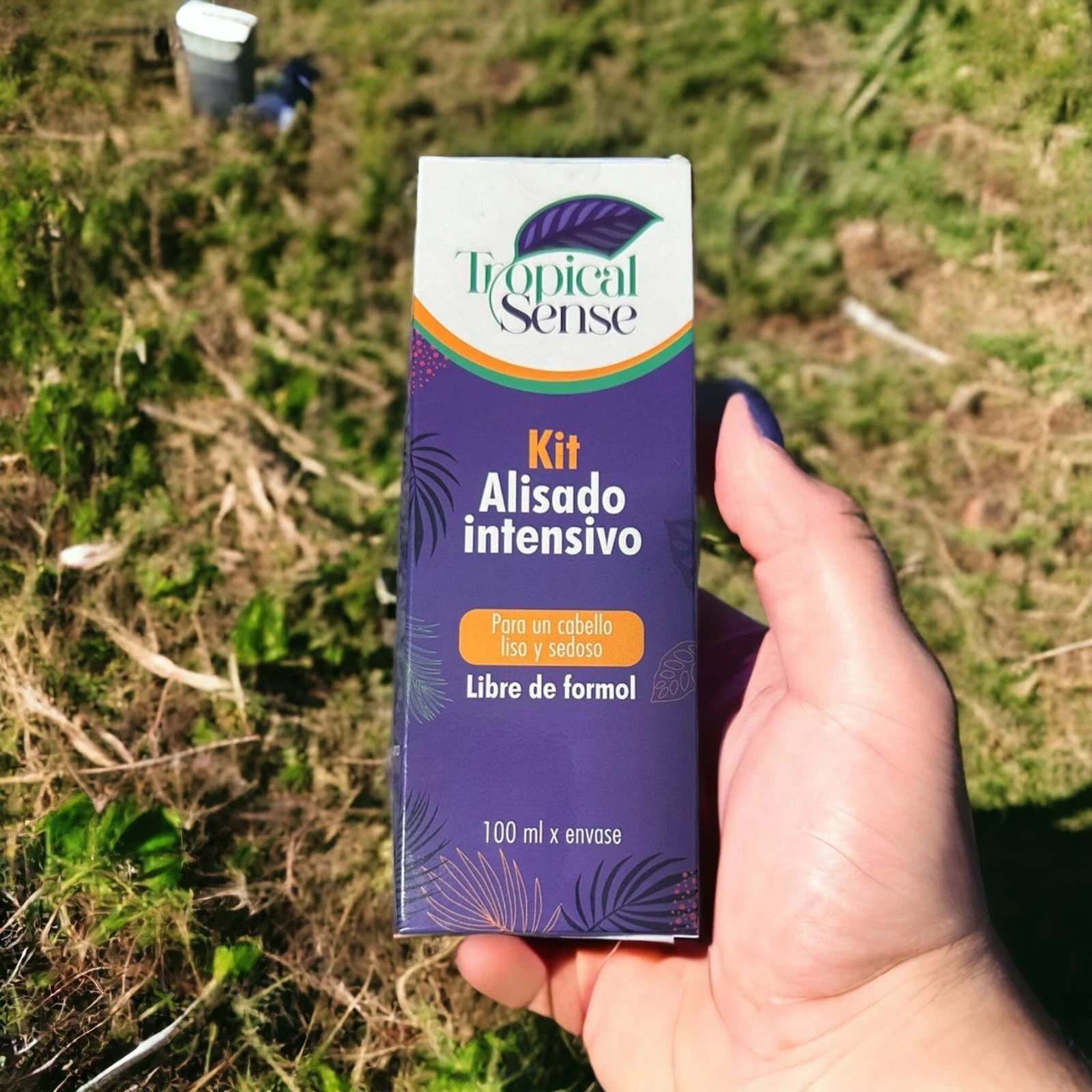 Mano sosteniendo caja de kit de alisado intensivo Tropical Sense, presentación de 100 ml, empaque morado con diseño de hojas sobre fondo de jardín.