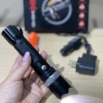 Linterna táctica negra, cuerpo metálico estriado, activada, con foco encendido y accesorios de carga al fondo.