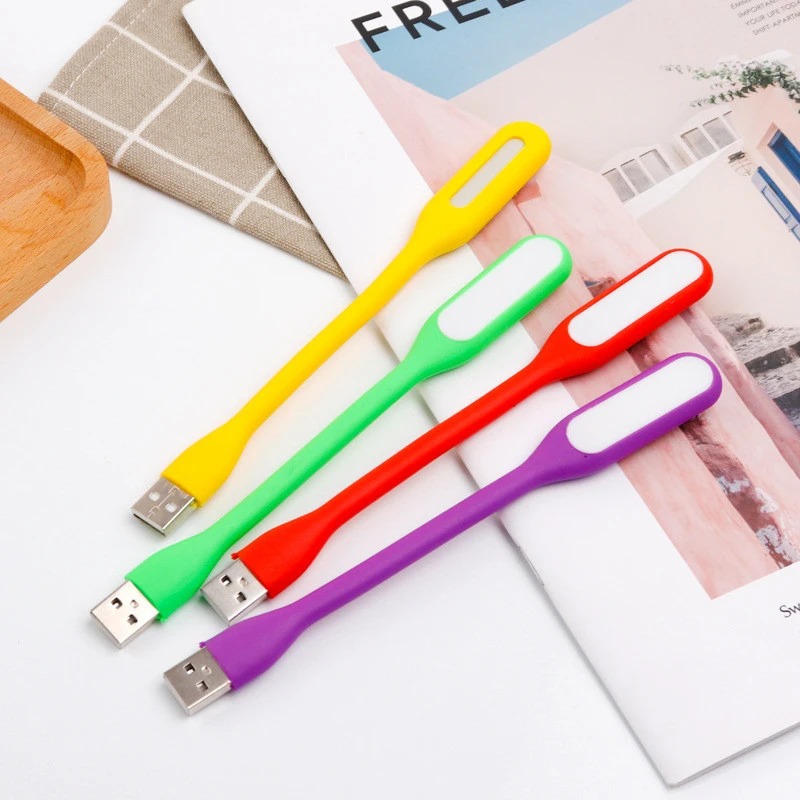Cuatro lámparas LED portátiles USB de silicona flexible en colores amarillo, verde, rojo y morado, dispuestas en diagonal sobre una revista abierta.