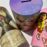 Tres alcancías cilíndricas: una con diseño de vino y firma Susan Winget, una floral con tapa morada, y una rosa de personaje infantil.